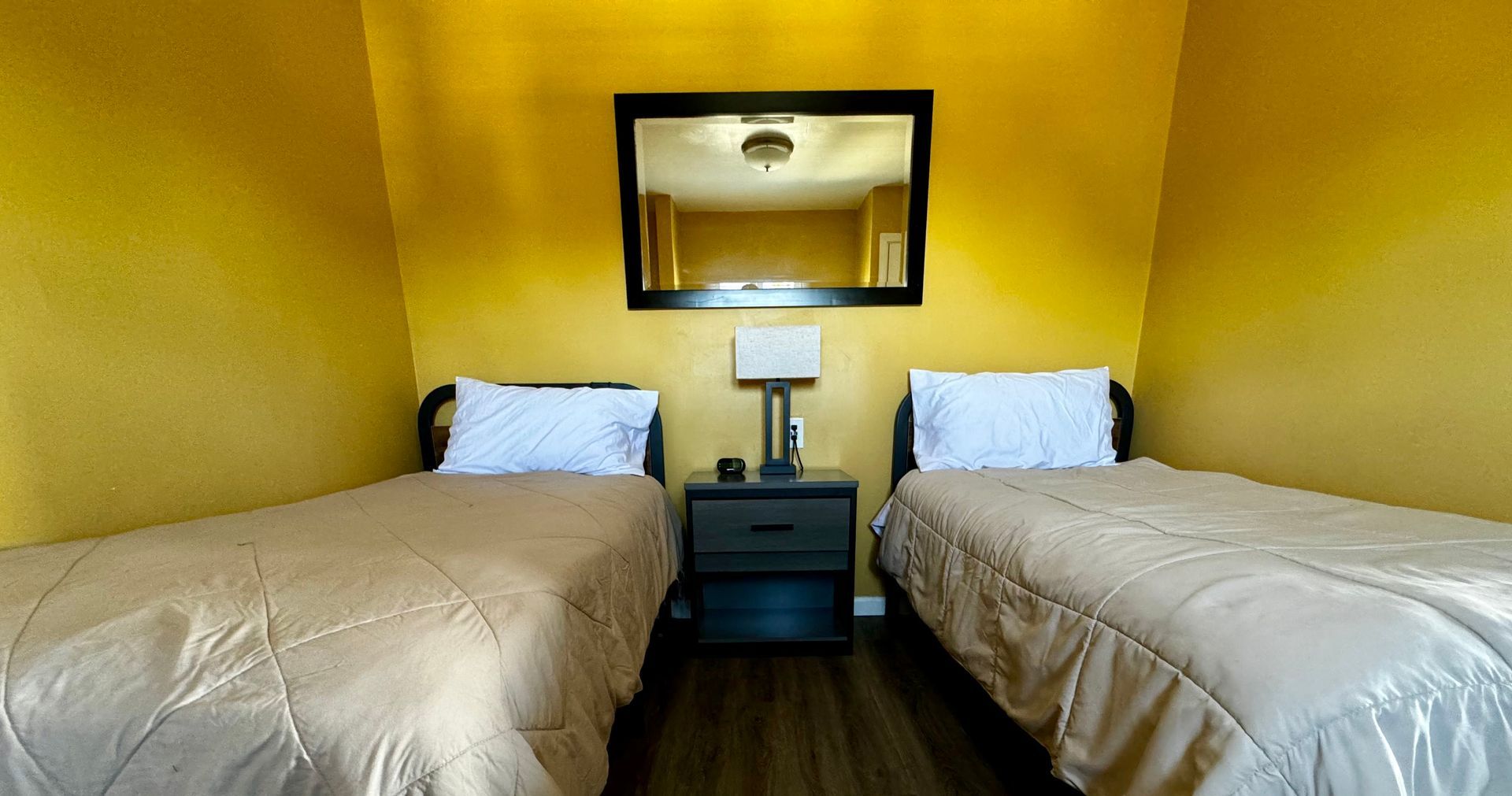 Two twin beds with beige comforters face a nightstand under a rectangular mirror against bright yellow walls.