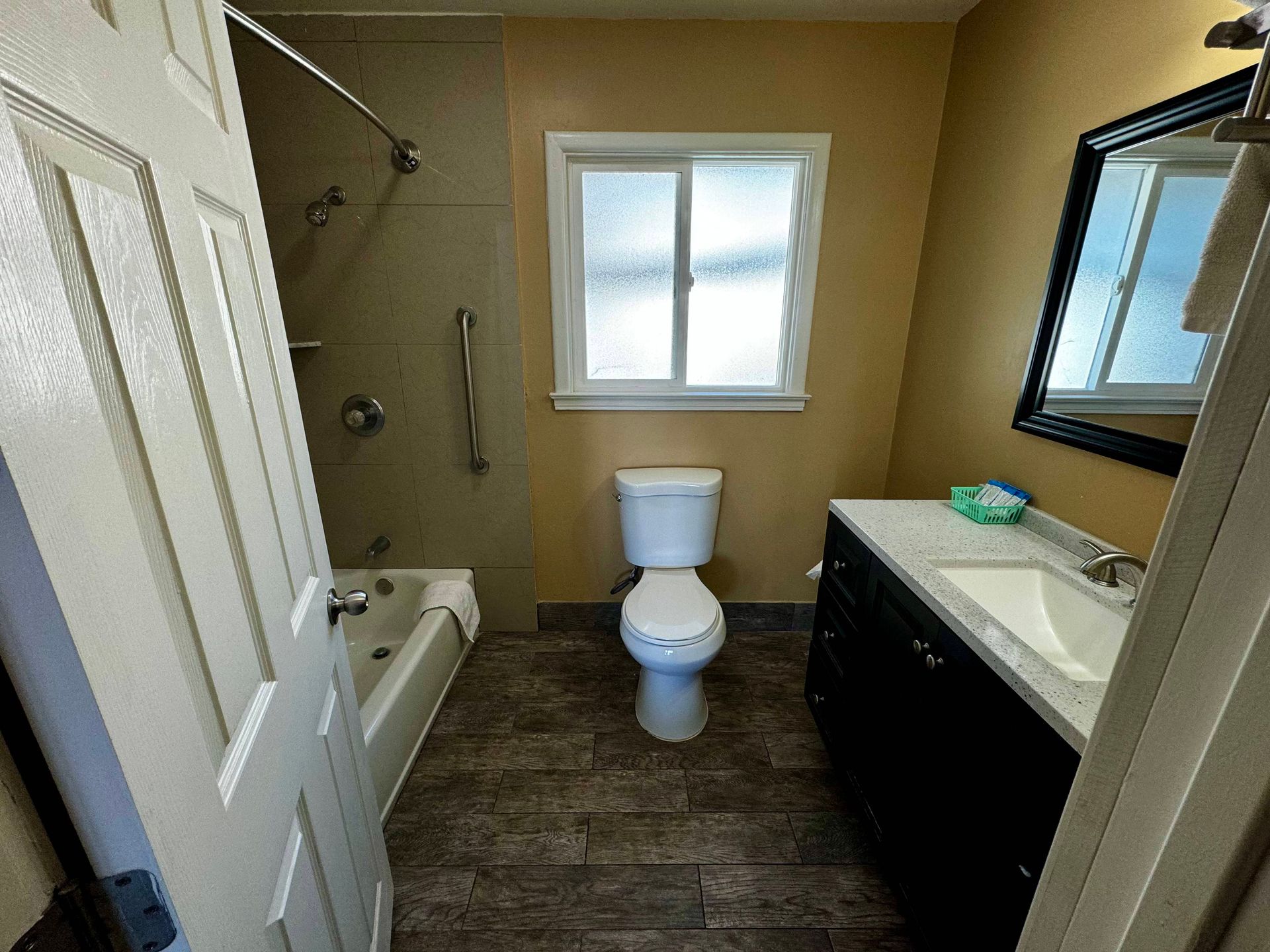 A bathroom featuring a white toilet, a bathtub with a shower head and grab bars, and a dark vanity cabinet with a mirror.