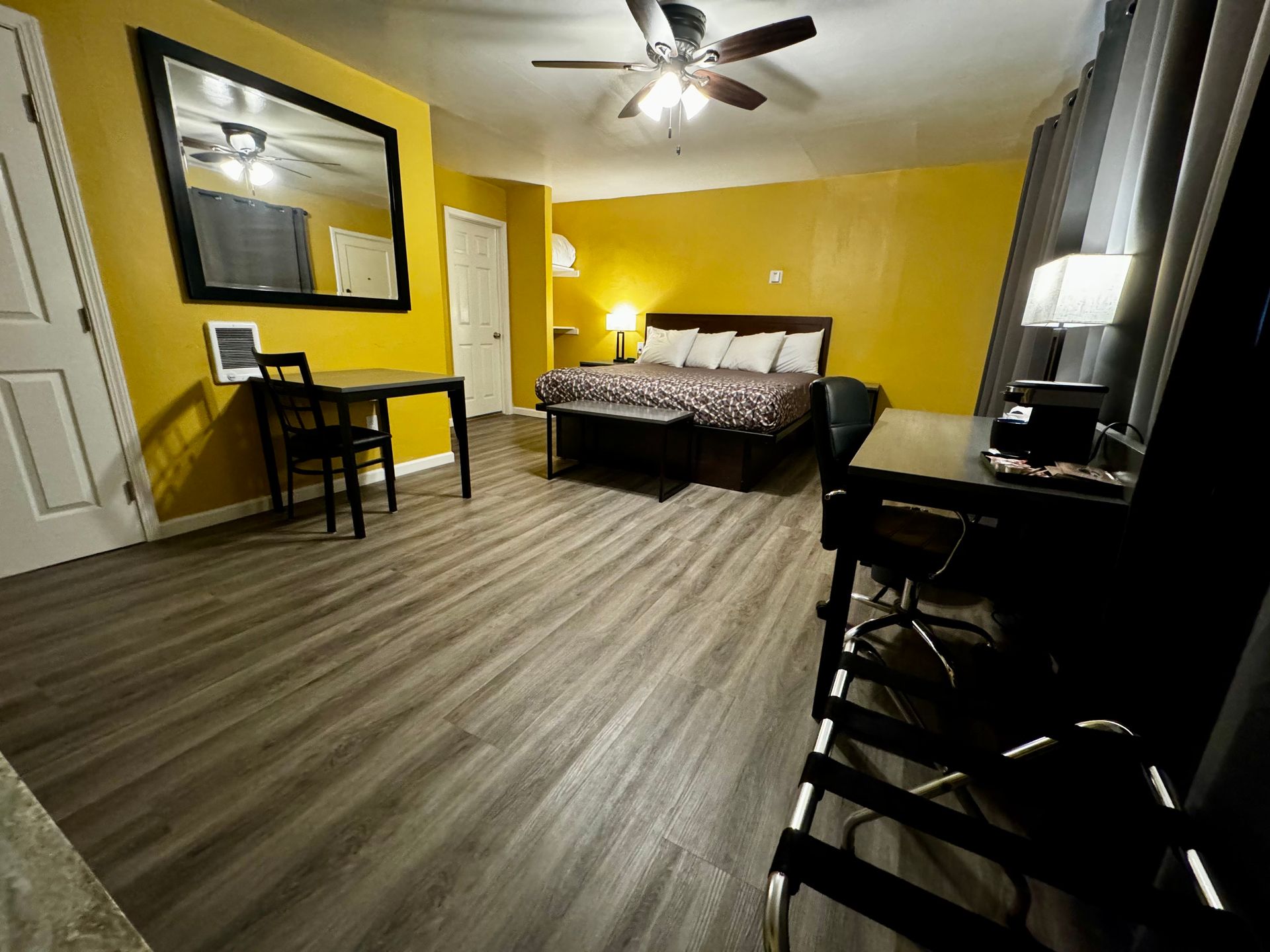 A hotel room with yellow walls, a bed, a small table with a chair, a desk area, and wood-grain flooring.