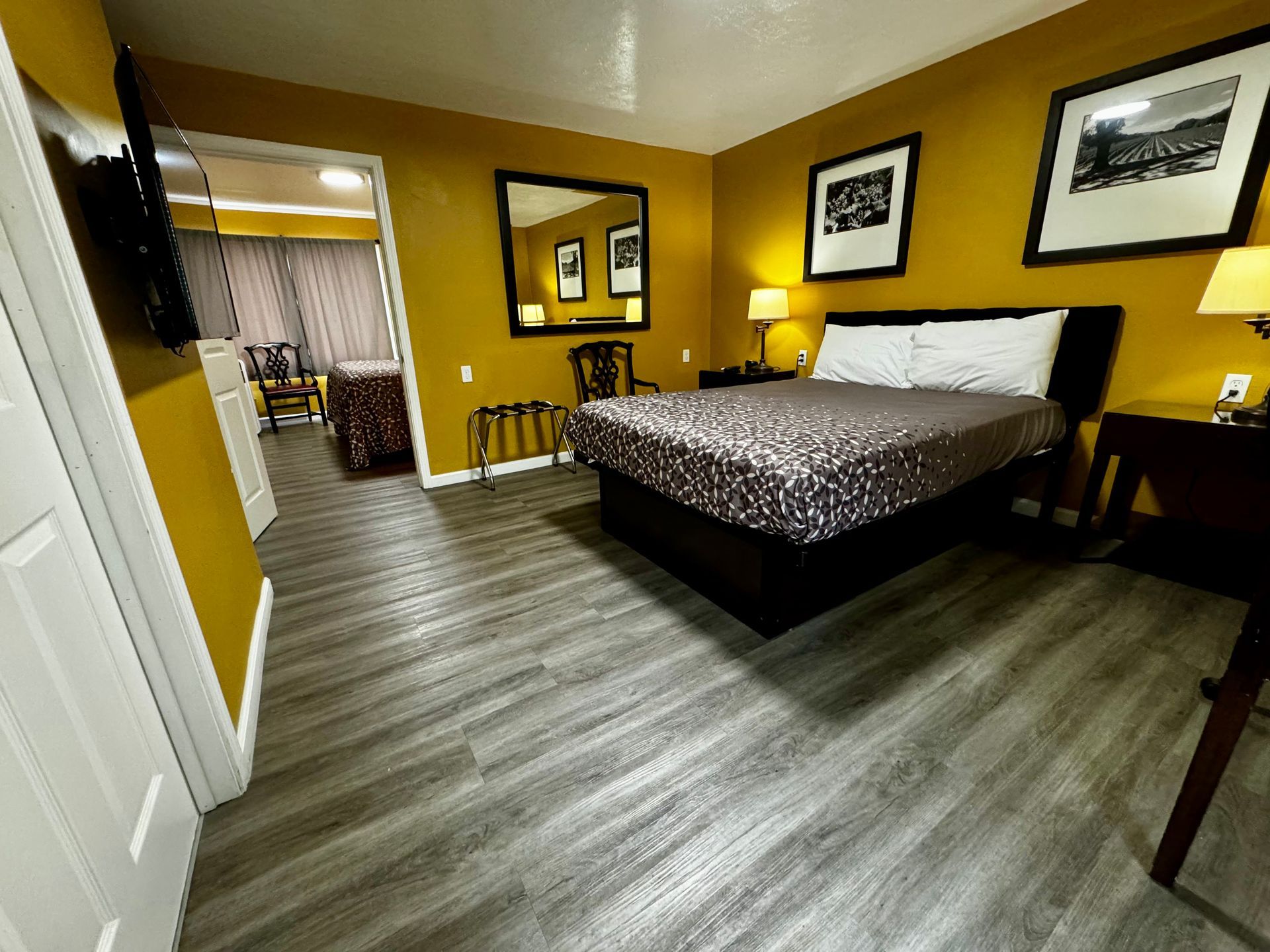 A hotel room with wood-look flooring, yellow walls, a double bed with a dark patterned comforter, and a view into a foyer.