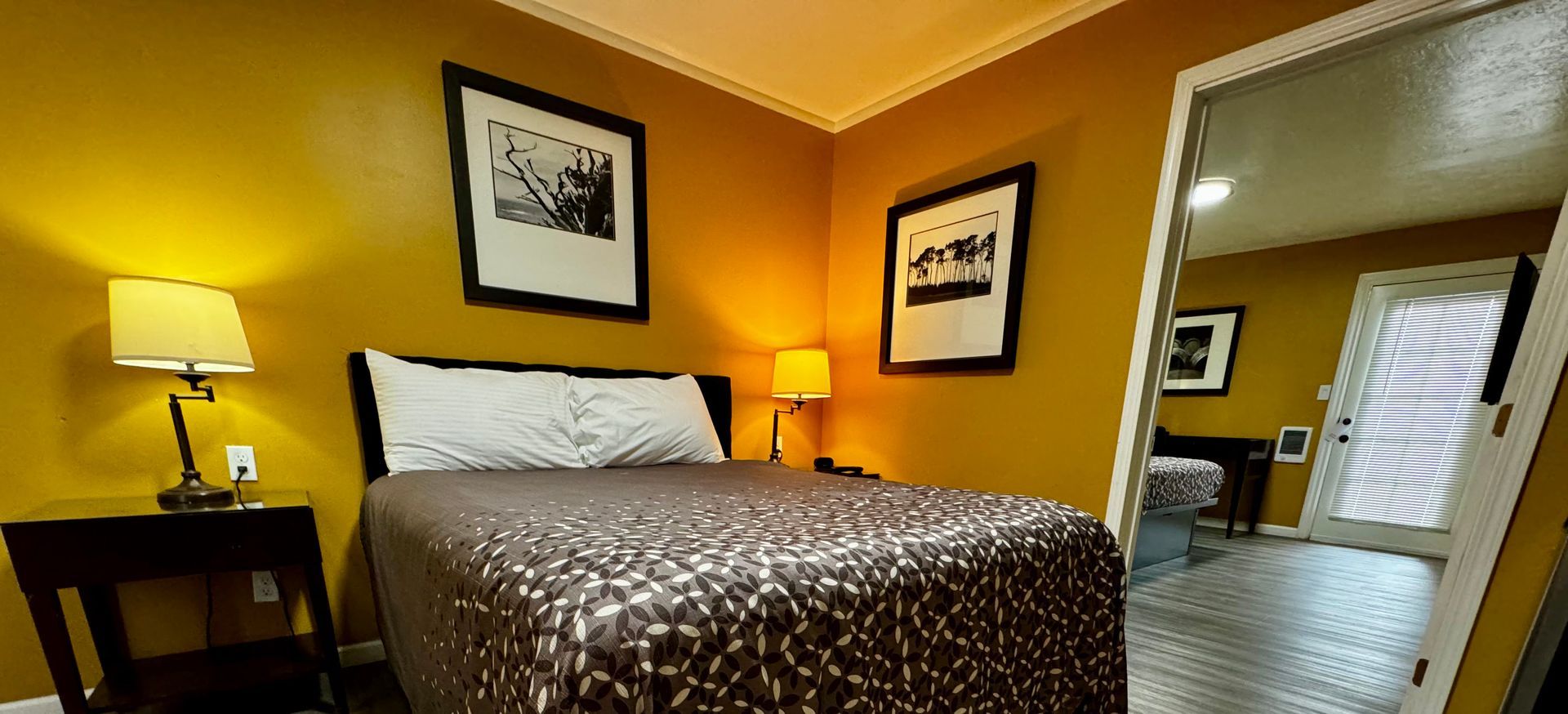A bedroom with a bed, a nightstand with a lamp, and wall art, viewed through an open doorway into a bright hallway.