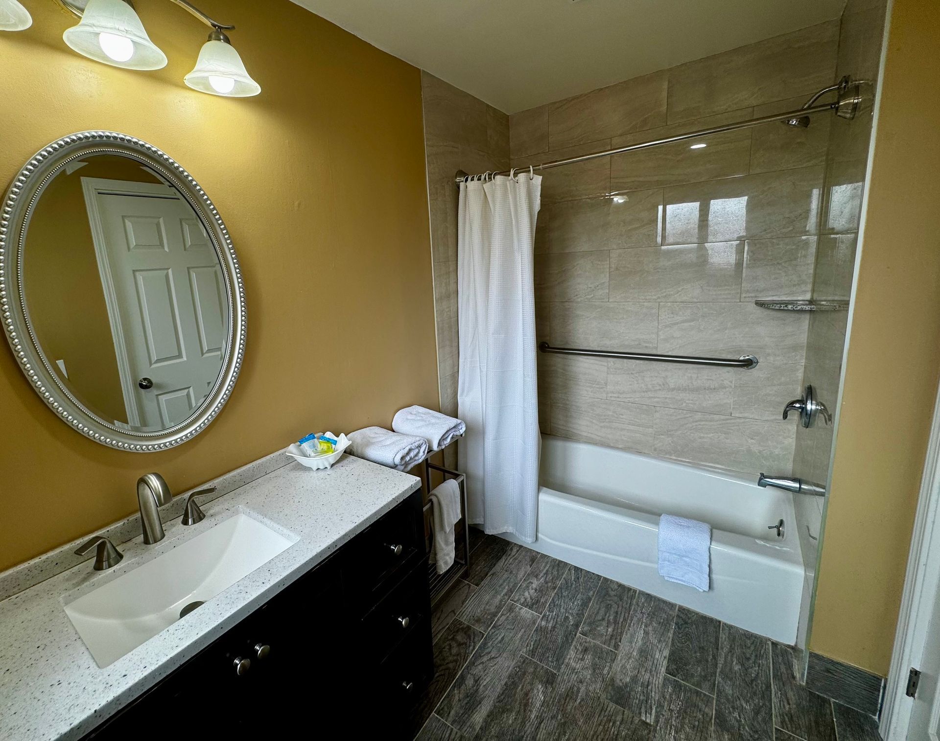 A bathroom with a dark vanity, oval mirror, white sink, and a tiled shower-tub combination with a white curtain.