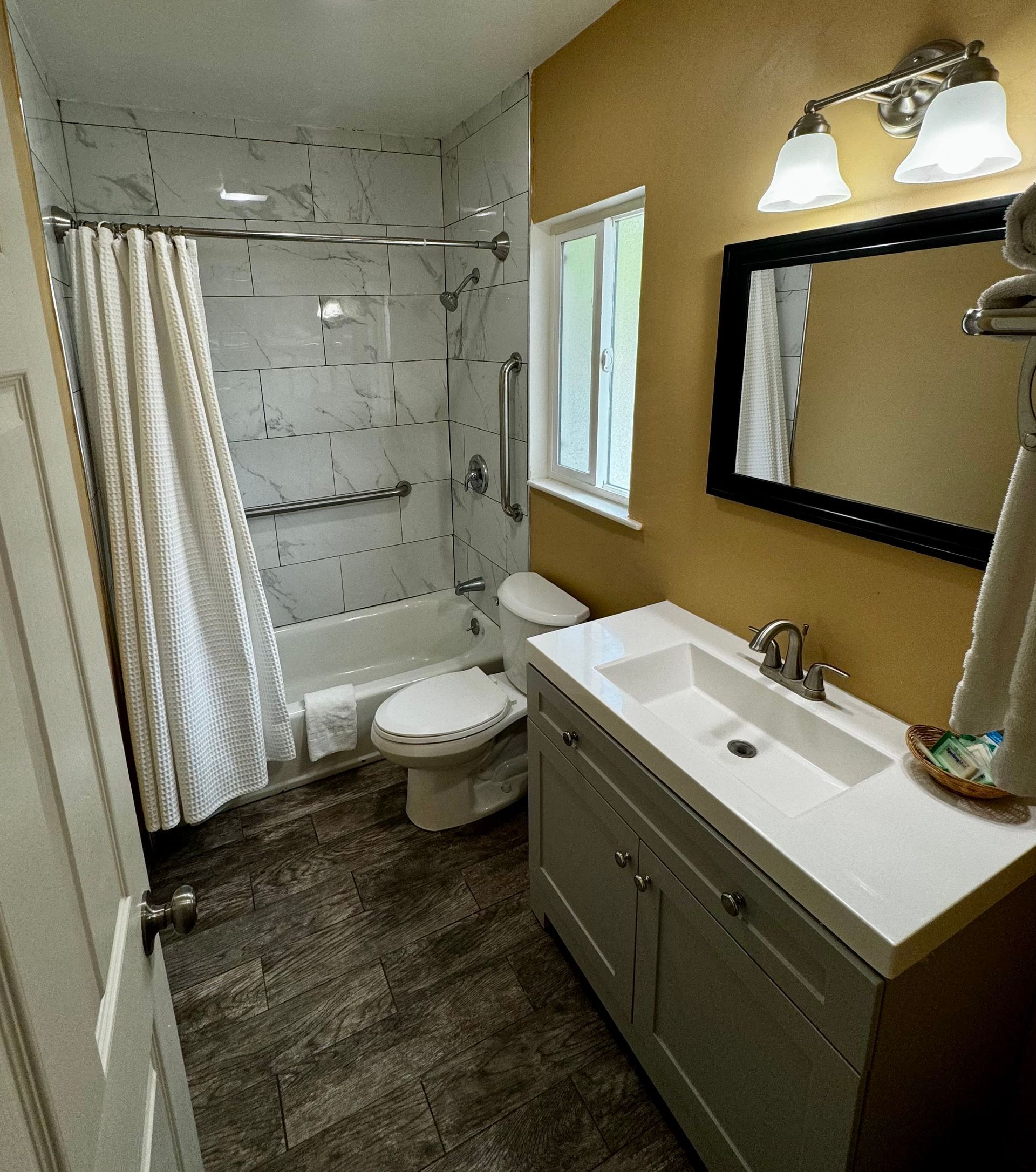 A small bathroom with a gray vanity, a sink, a mirror, a toilet, and a tiled shower with a curtain against gold walls.