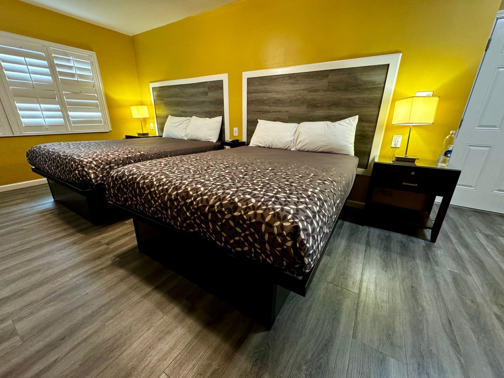 A hotel room with two queen beds featuring dark patterned bedding, wooden headboards, and a yellow accent wall.