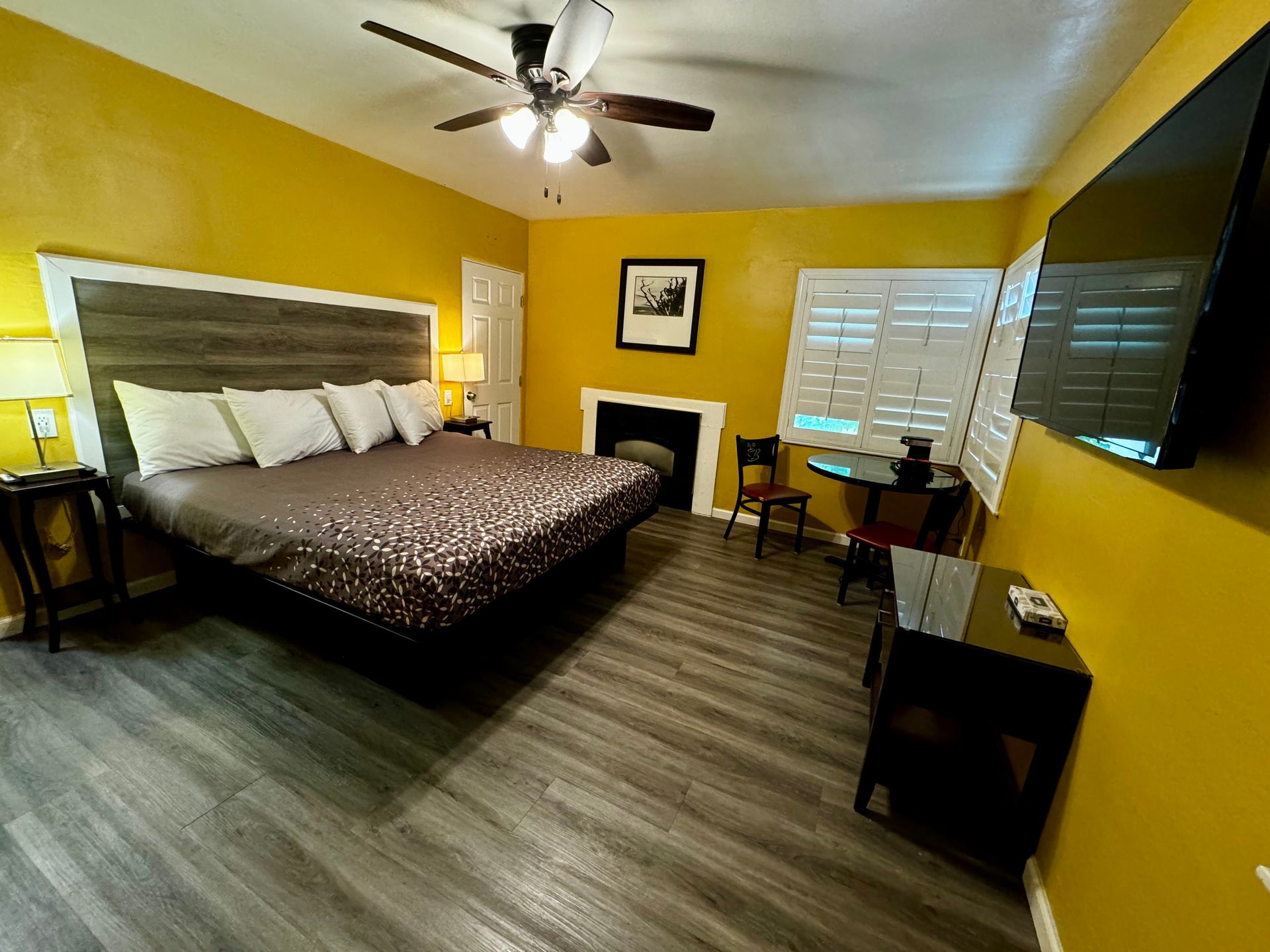 A bright yellow bedroom with a king-size bed, gray wood-look flooring, a wall-mounted TV, and a small dining set.