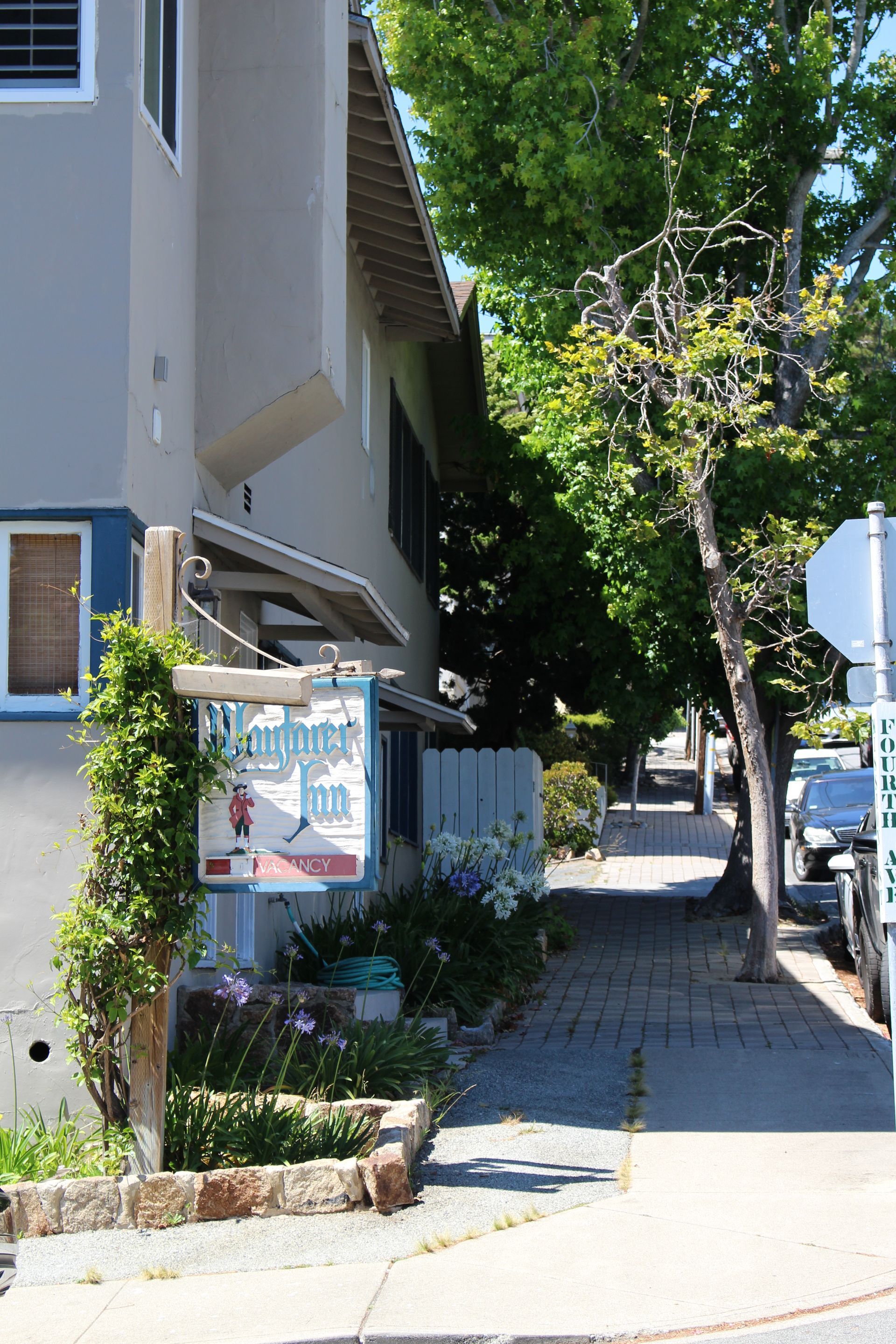 A view down a sidewalk along a light-colored building with a hanging sign, green trees, and parked cars in the distance.