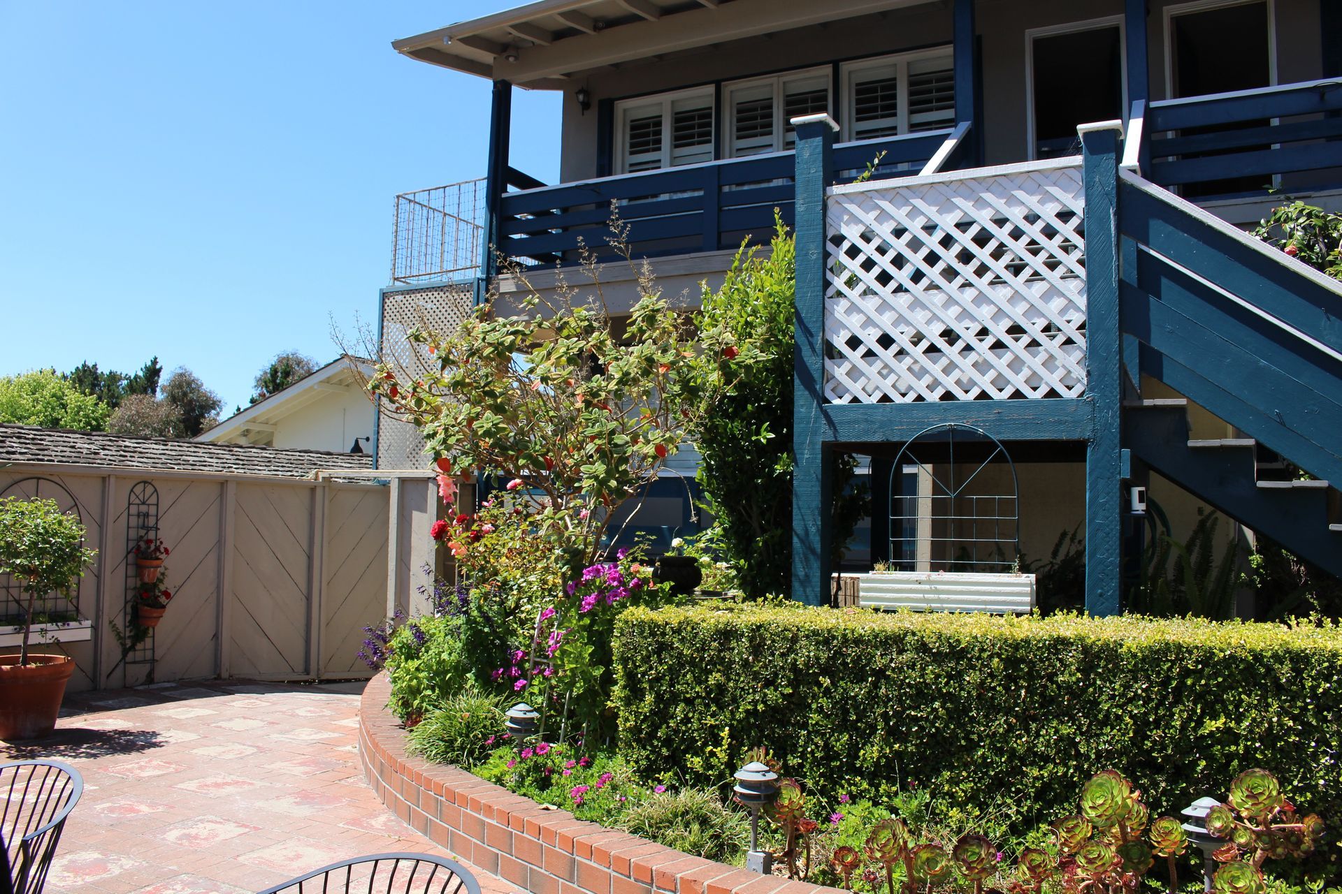 A multi-story building with a blue deck, white lattice, and stairs, beside a landscaped garden with a brick-edged hedge.