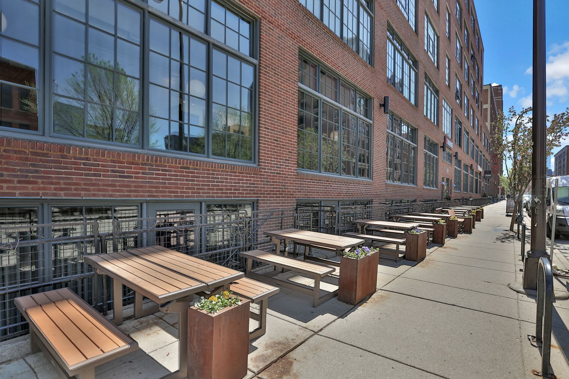 A row of wooden tables and benches on a sidewalk in front of a brick building.