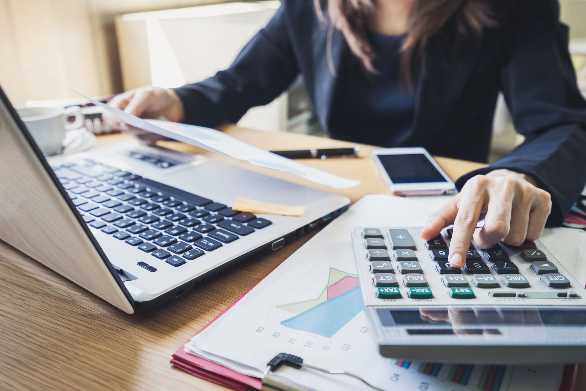 Two people reviewing financial documents, one using a calculator, the other pointing at laptop graphs.
