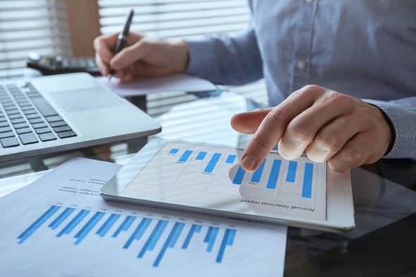 Person analyzing charts on tablet and paper, writing with a pen, laptop visible.