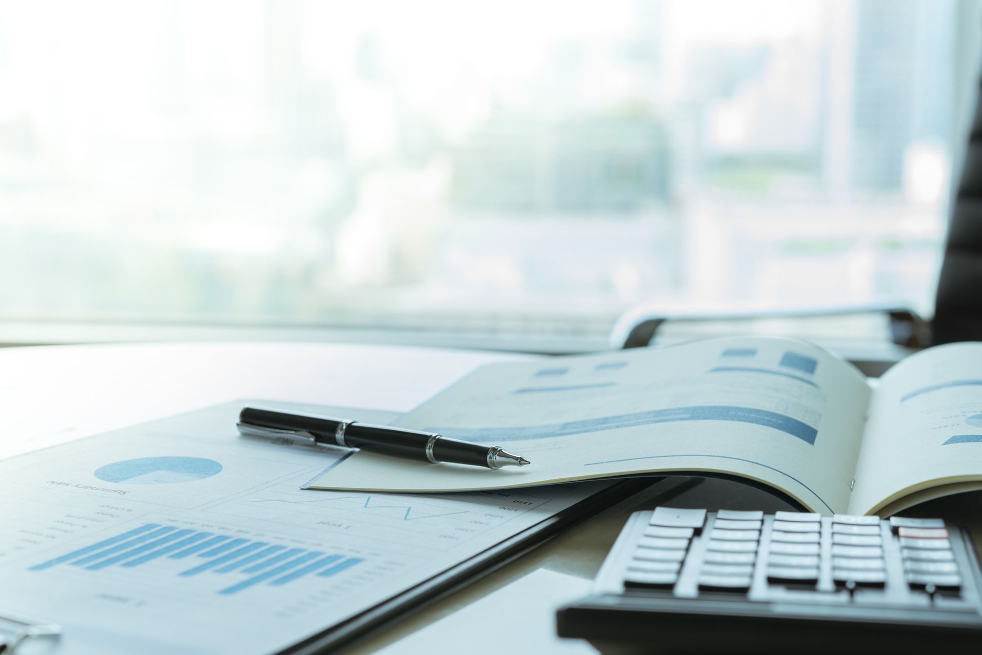 Desk with financial documents, pen, and calculator near a bright window.