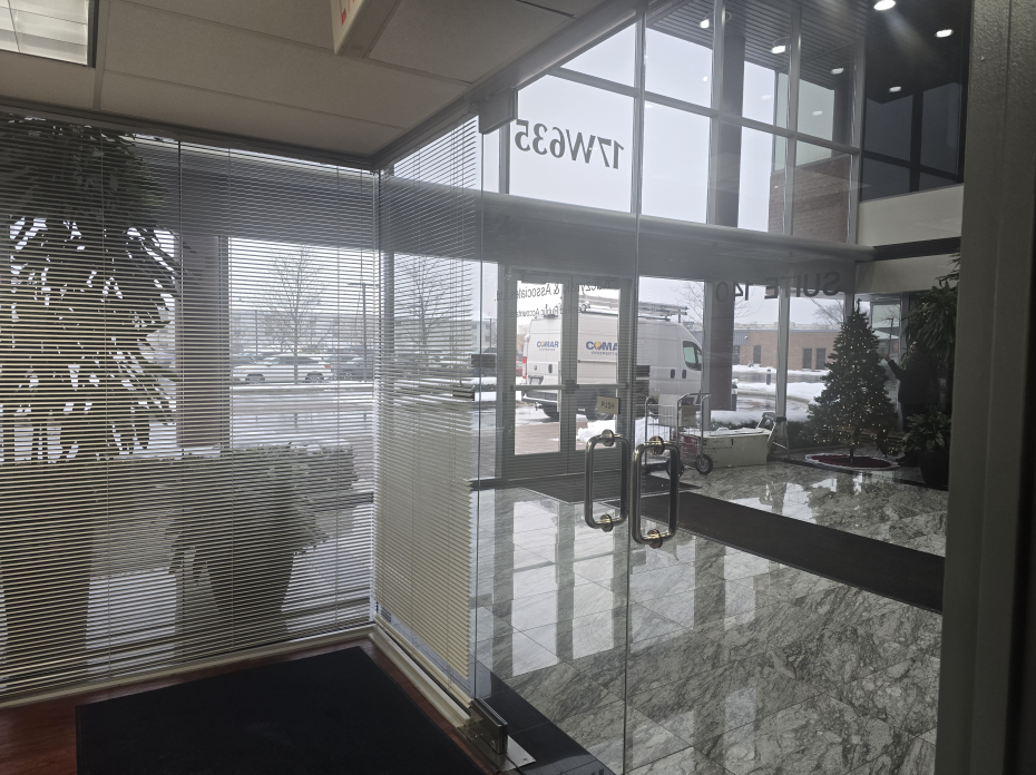 Close-up of a wooden table with a stack of papers, pens, and eyeglasses, with blurred chairs and window in the background.