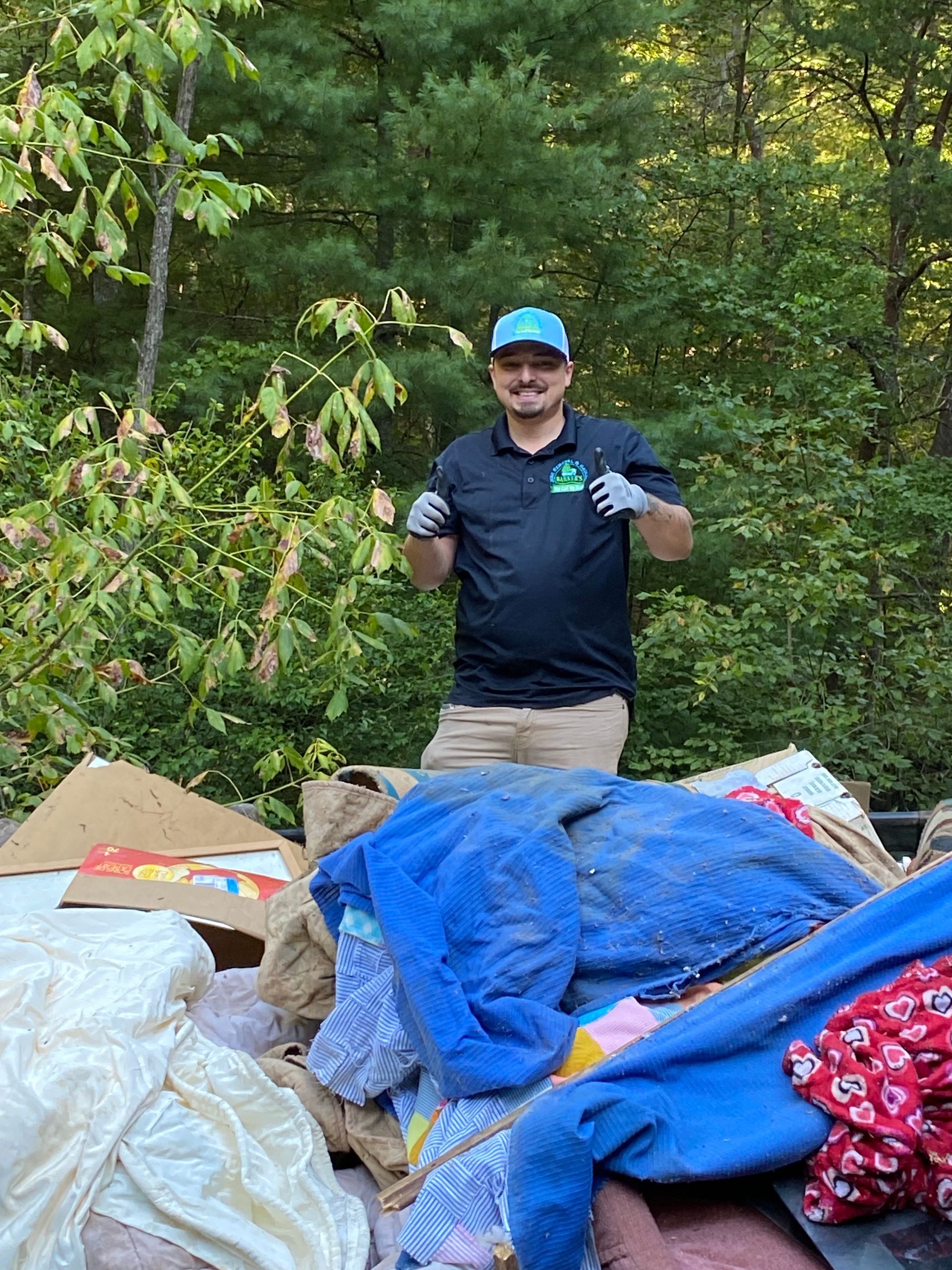 Man in black shirt and hat, smiling, standing by a pile of discarded clothes in an outdoor setting.