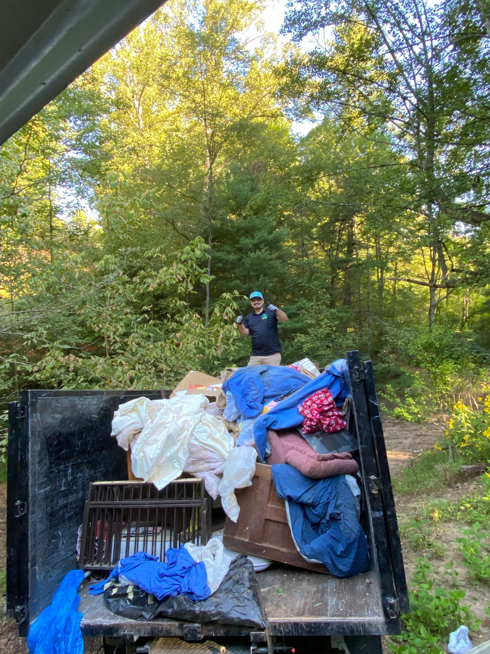 A person standing in a truck bed filled with discarded items, in a wooded area.