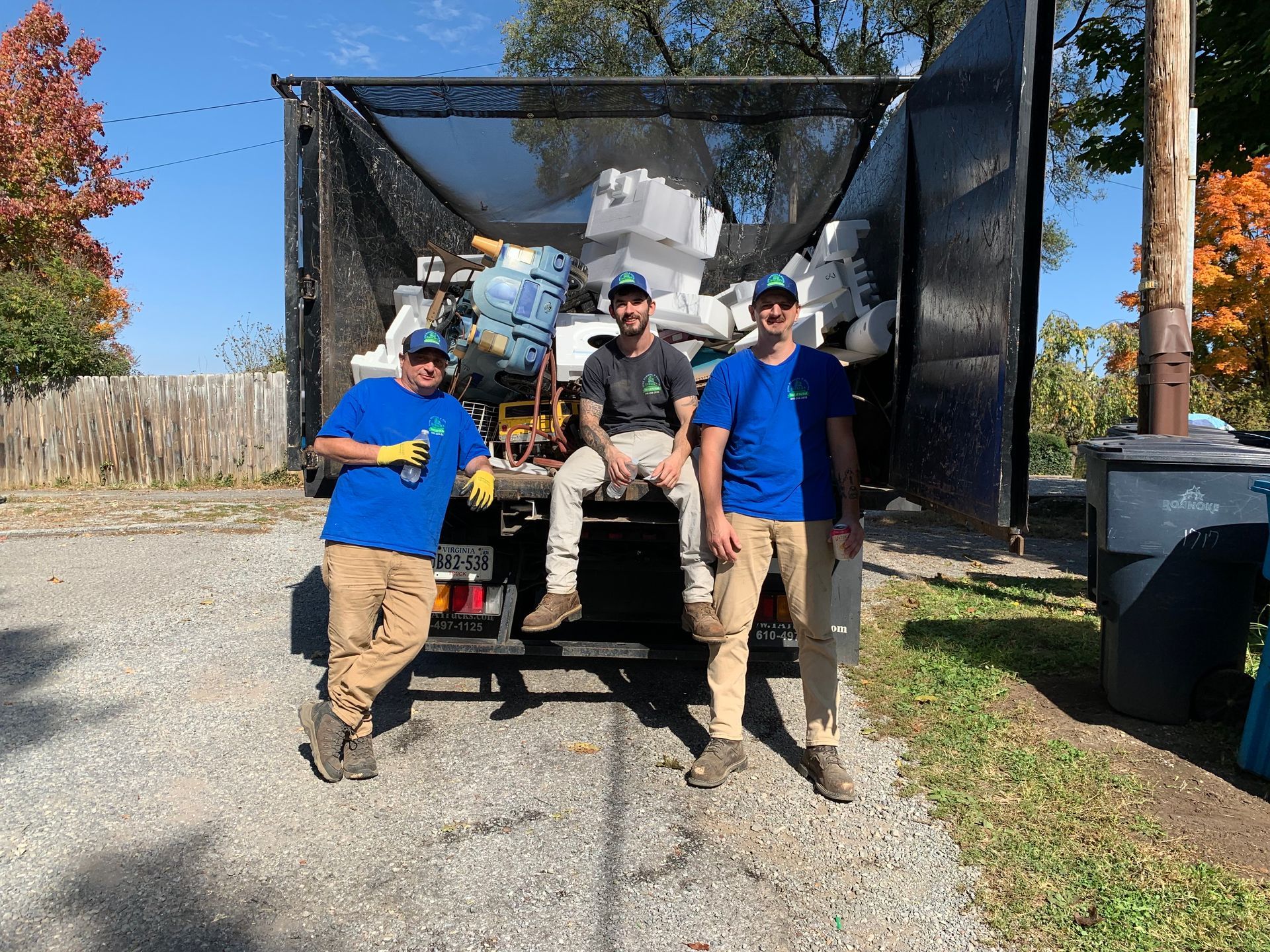 Three men in blue shirts stand by a truck filled with debris, smiling outdoors.