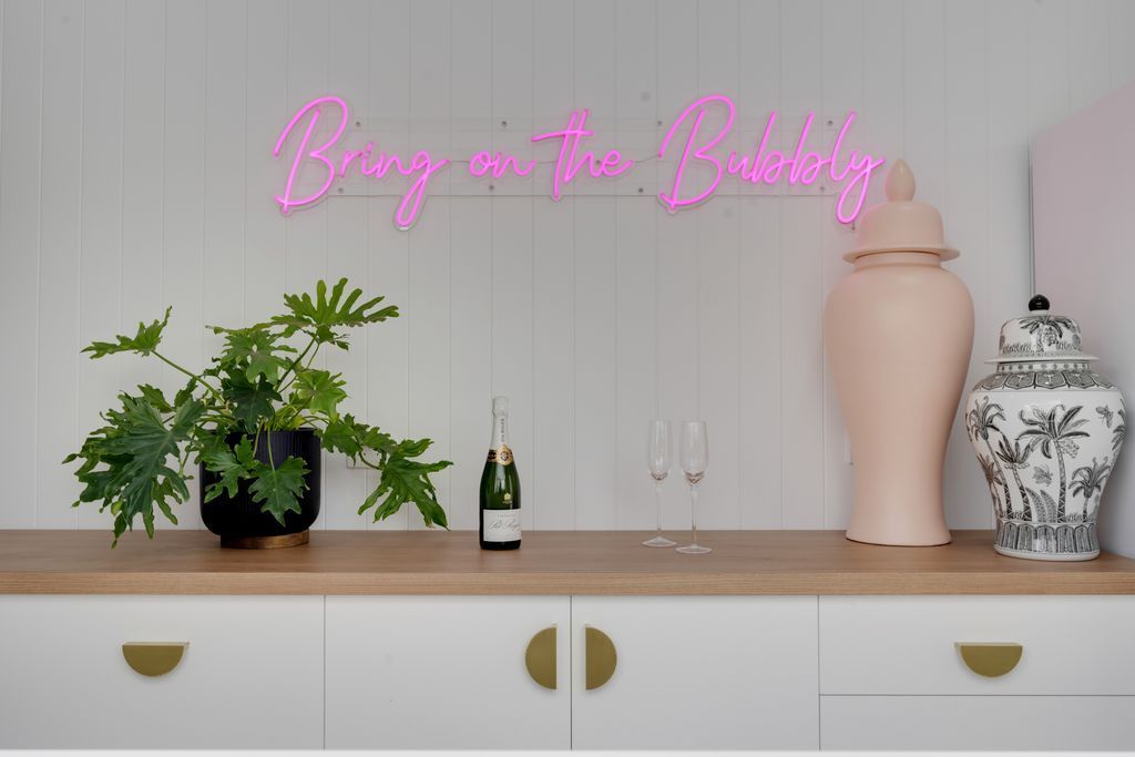 Pink neon “Bags on the Back” sign above a console with plants, vases, and white cabinetry