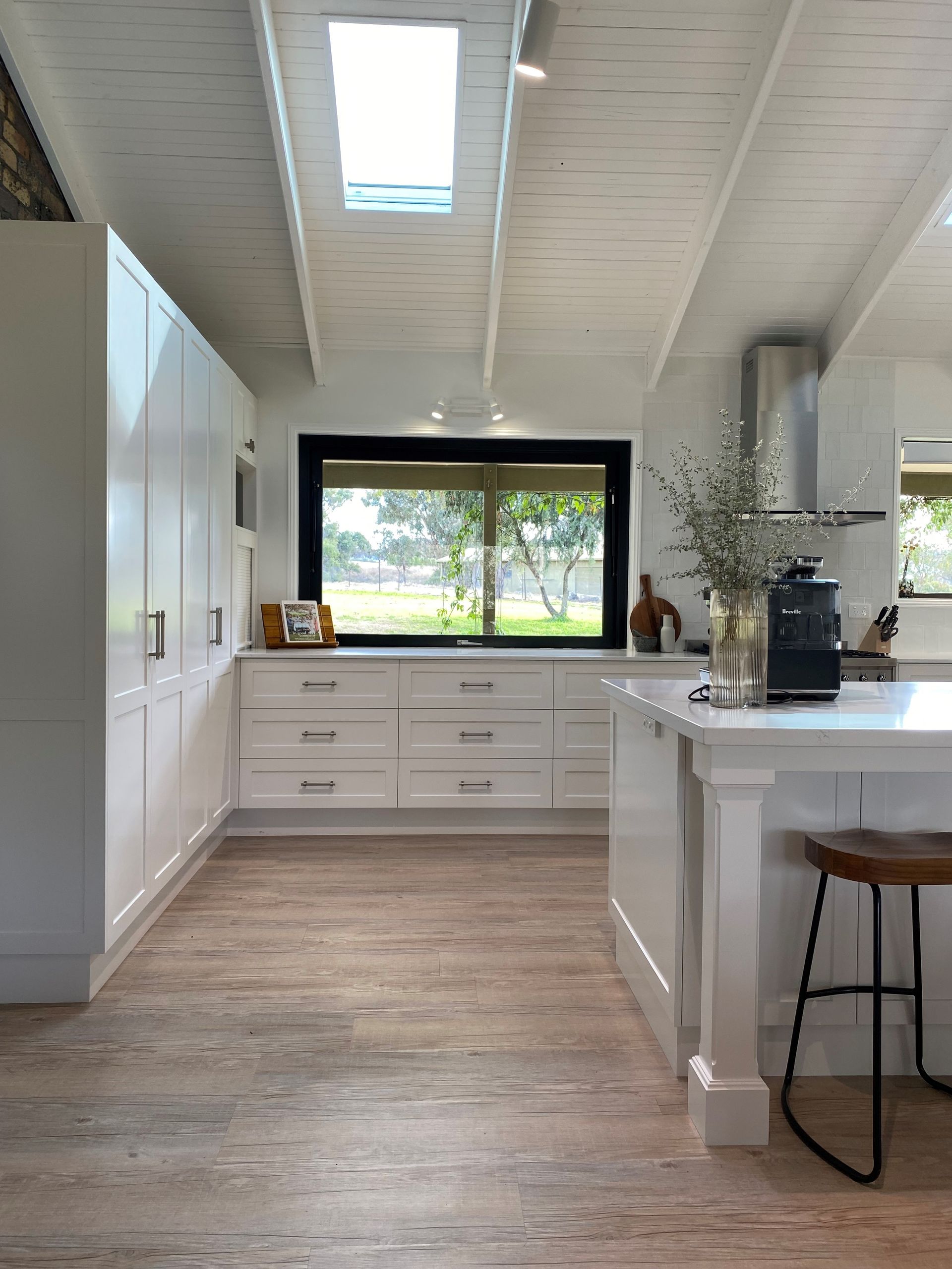 Bright white kitchen with wood floors, island seating, and a large window overlooking trees