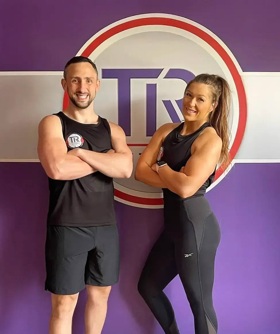 Two people posing in front of a logo. Man and woman in workout clothes with arms crossed, smiling. Purple and white background.
