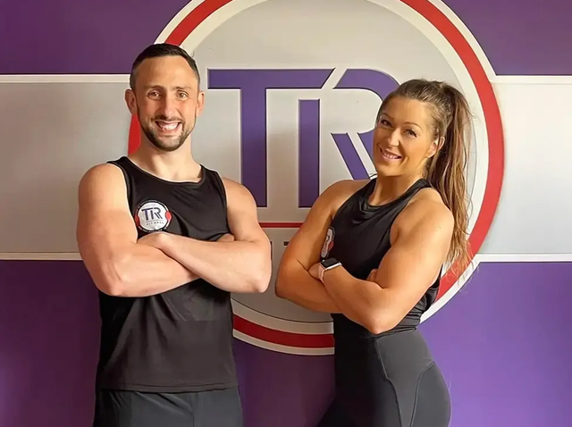 Two people posing in front of a logo. Man and woman in workout clothes with arms crossed, smiling. Purple and white background.
