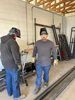 Two men are standing next to each other in a garage wearing welding helmets.