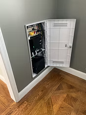 Wall-mounted white panel door open, revealing black electronic equipment, on gray wall, over parquet flooring.