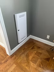 White electric baseboard heater on a gray wall in a corner of a room with parquet flooring.