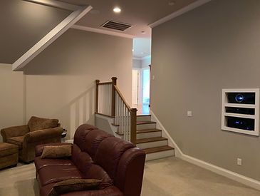 A home theater room with a brown leather couch, staircase, and AV equipment in the wall.