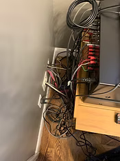 Tangled cables and wires behind a wooden cabinet, near a wall with electrical outlets, and a wooden floor.