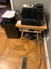 Wooden table with electronics, cords, and a printer on a patterned wood floor near a wall with a vent.