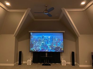Home theater with projector screen, speakers, and a ceiling fan, set in a room with angled walls and recessed lighting.