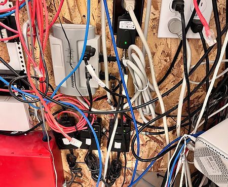 A tangled mess of electrical cords, power strips, and connected devices against a wood-paneled wall.