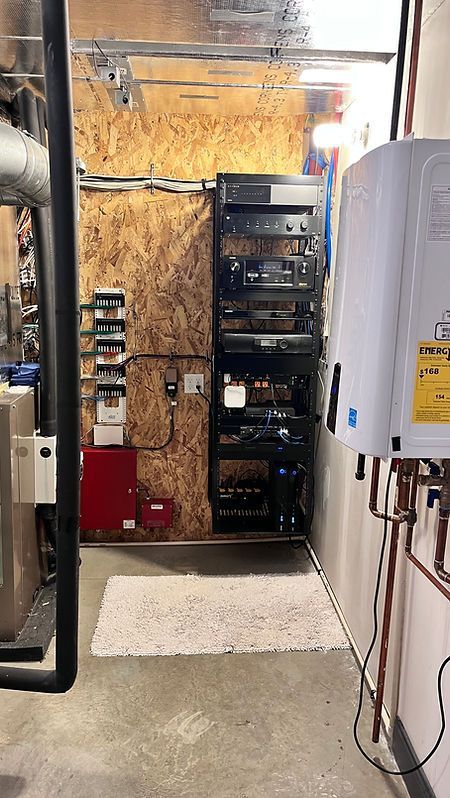 A utility room with a black equipment rack, water heater, and electrical panels against wooden and concrete walls.