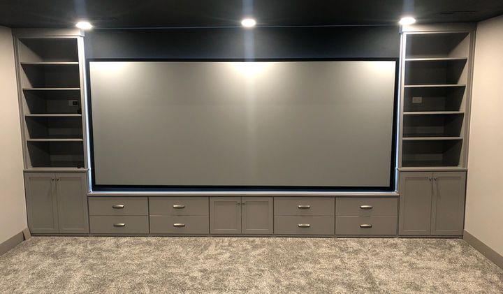 Built-in entertainment center with a large screen, shelves, drawers, and cabinets; gray with dark ceiling.
