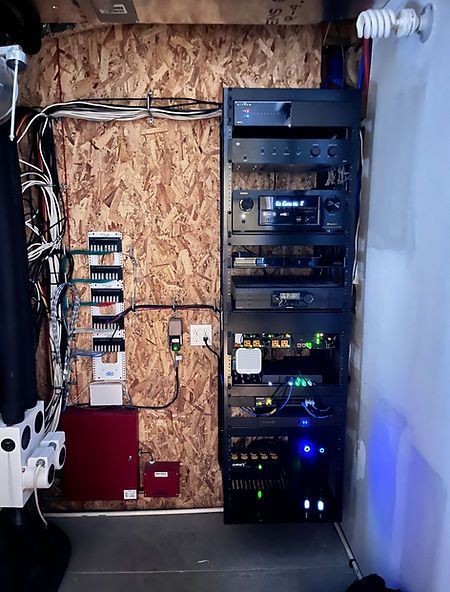 A home theater equipment rack with various black components against a wood-paneled wall, cables visible.