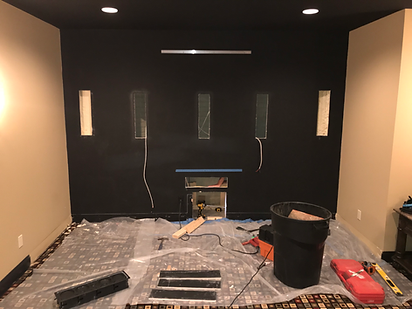 Black wall with in-wall speaker cutouts, wires, and construction materials on the floor of a home theater.