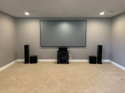 Home theater room with large screen, speakers, equipment stand, and beige carpet.
