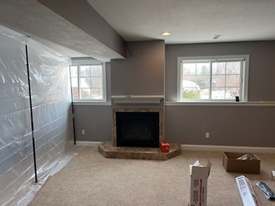 Basement room with a fireplace and two windows, painted gray, plastic sheeting on one wall, boxes on the floor.