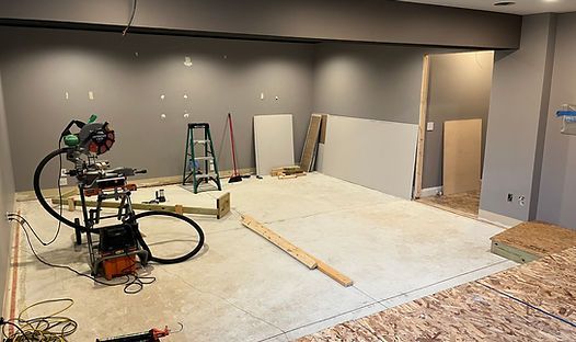 Room under construction with tools, drywall, and partially installed flooring; grey walls, bare concrete floor.