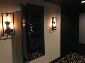 Wall-mounted media cabinet with open door, revealing electronic components, flanked by sconces, in a home theater setting.