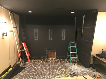 Room under construction, black wall with speaker cutouts, ladders, floor partially covered, beige side wall.