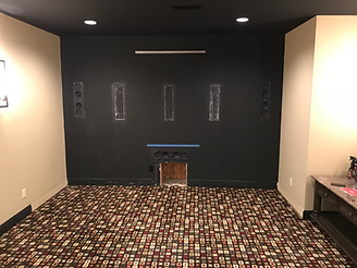 Black accent wall with speaker outlines in a home theater. Beige walls, black ceiling and patterned carpet.