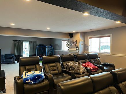 A home theater with black leather recliners, a small trampoline visible through a doorway, and recessed ceiling lights.