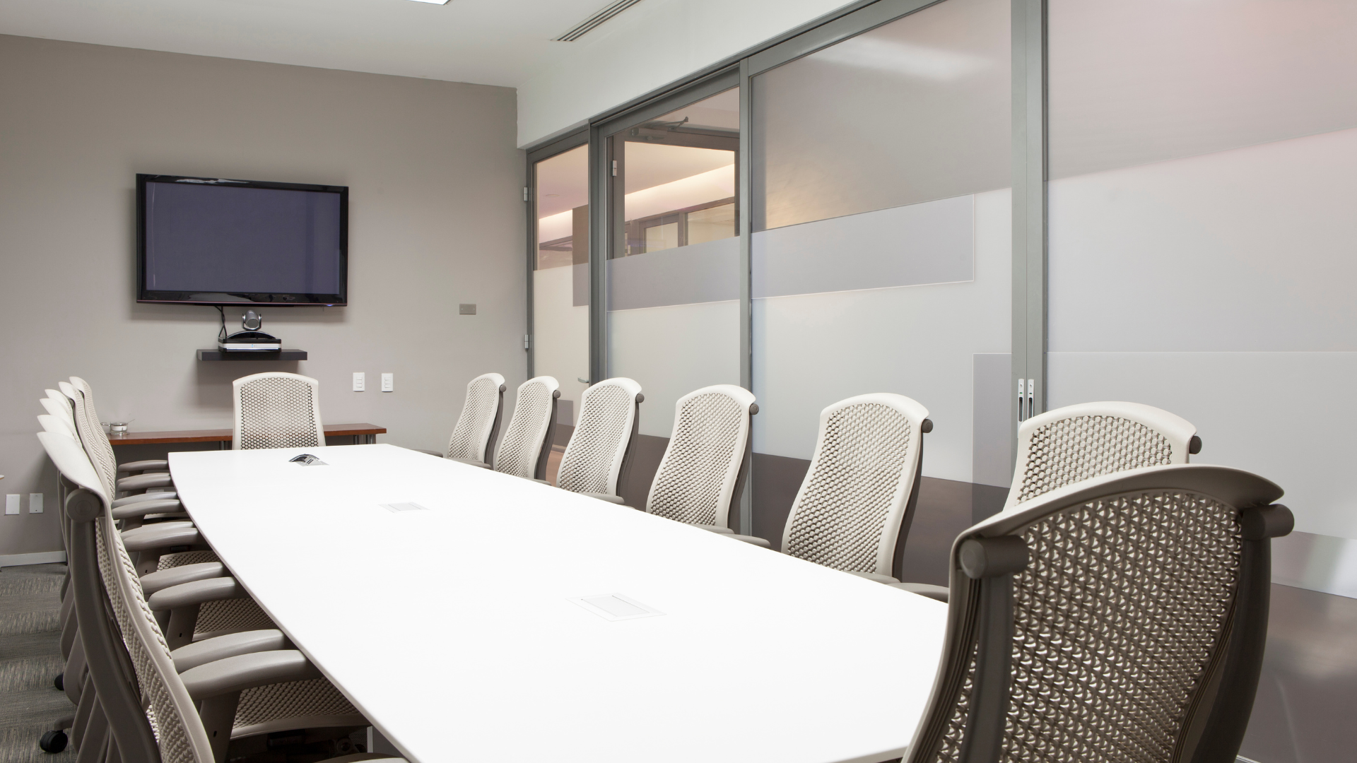 Conference room with long white table, chairs, and mounted TV. Frosted glass wall.