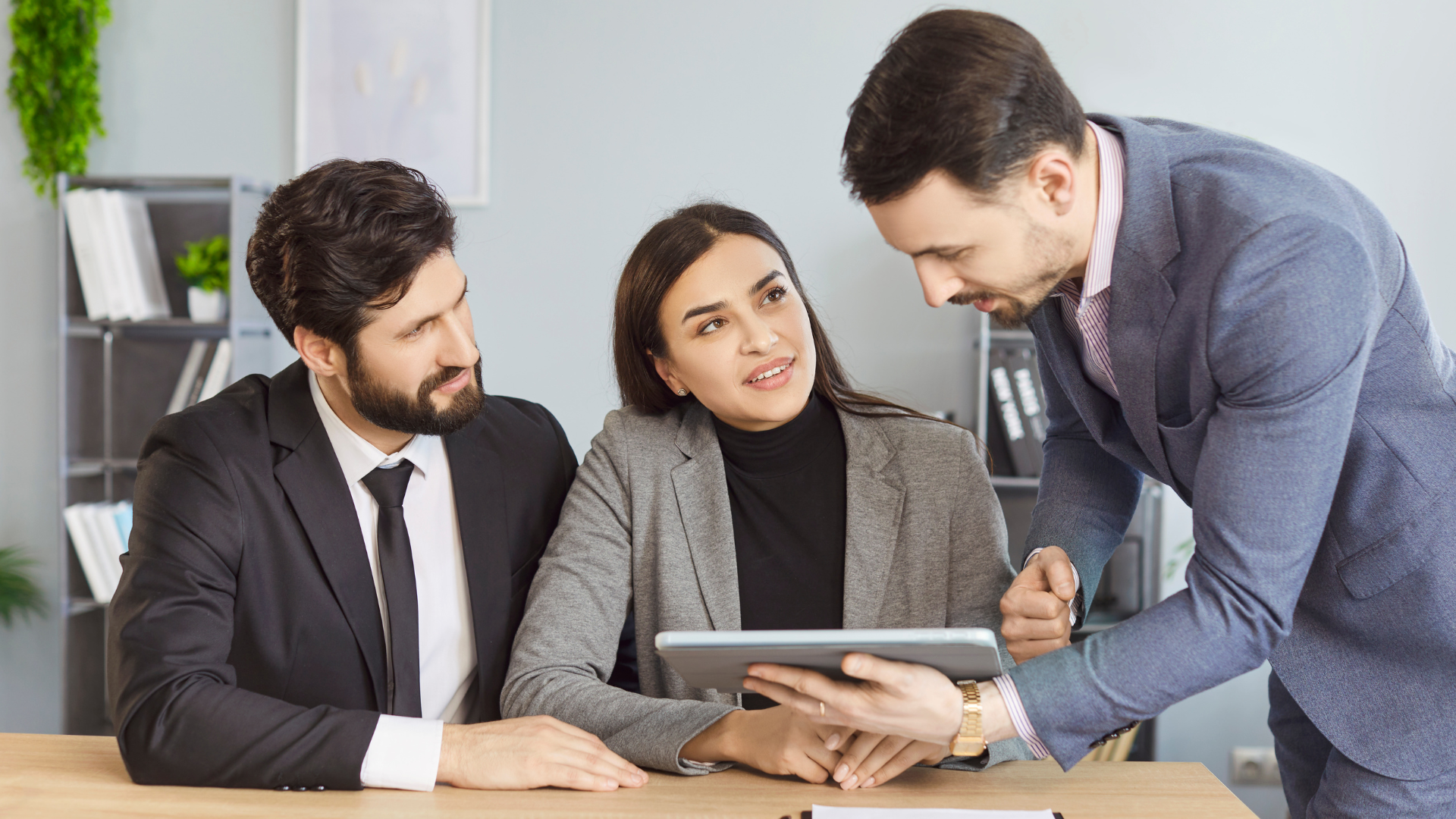 Three people in business attire reviewing a tablet in an office setting.