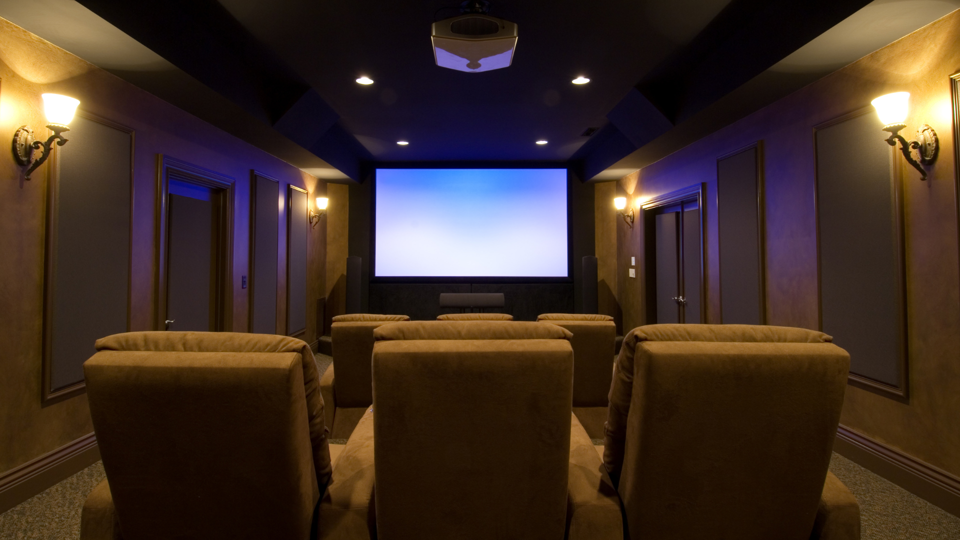 Home theater with tan recliners facing a large screen. Dim lighting, sconces, and a ceiling projector.