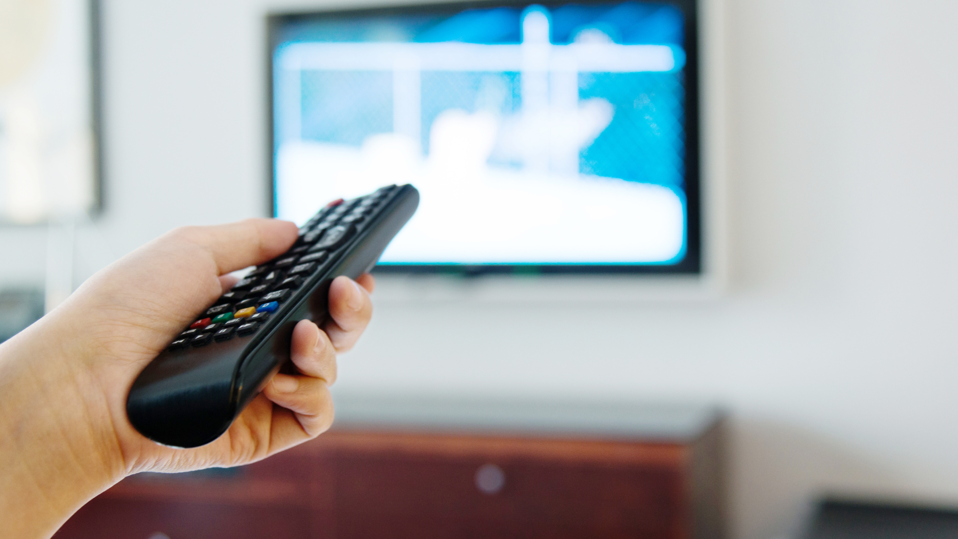 Hand holding a black remote control, pointing at a blurred TV screen.