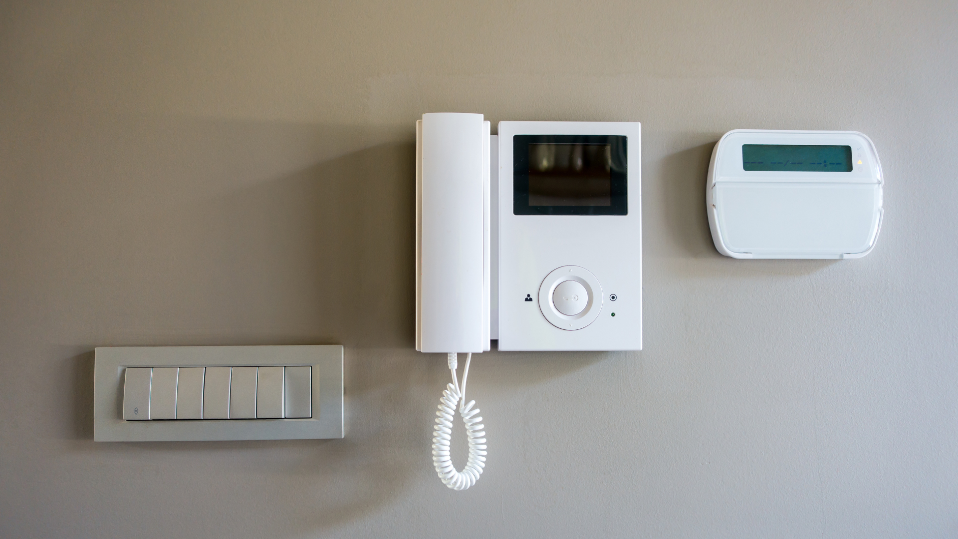 White intercom, thermostat, and light switches mounted on a beige wall.