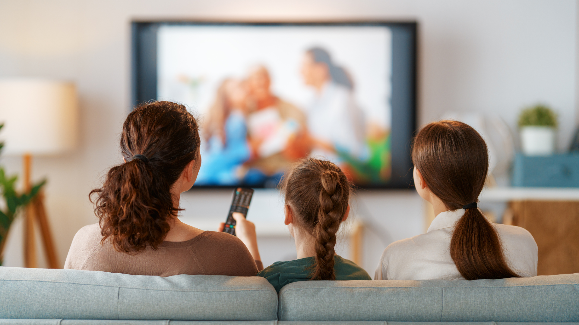 Three people sit on a couch watching TV; one holds a remote. Interior setting.