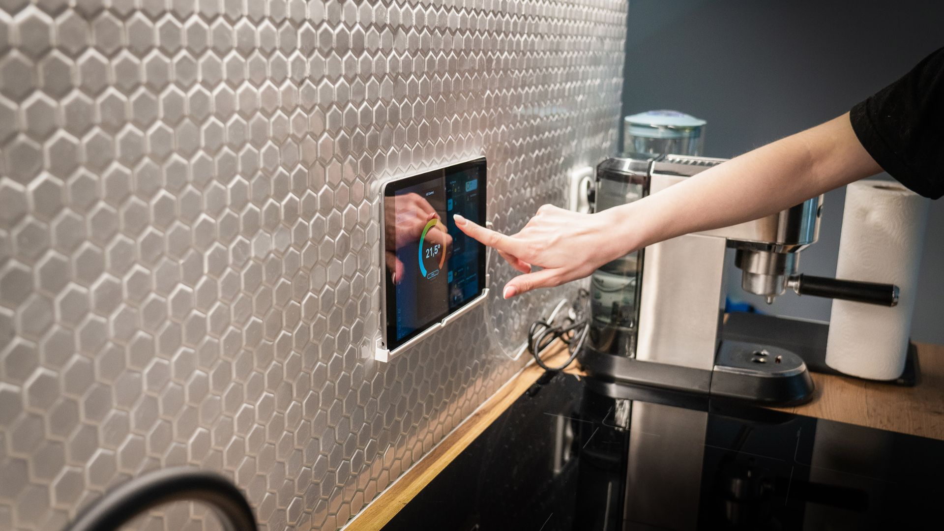 Hand touches a tablet embedded in a tiled wall in a kitchen. A coffee machine is in the background.