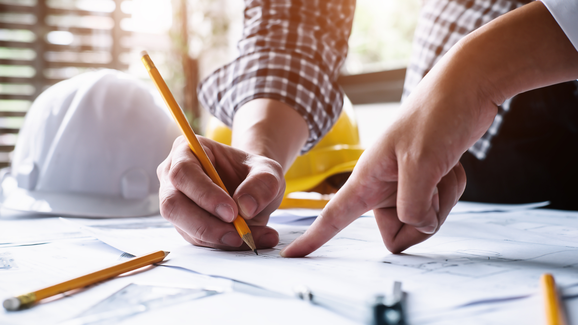 Person in plaid shirt points to construction plans while holding a pencil; two hard hats nearby.